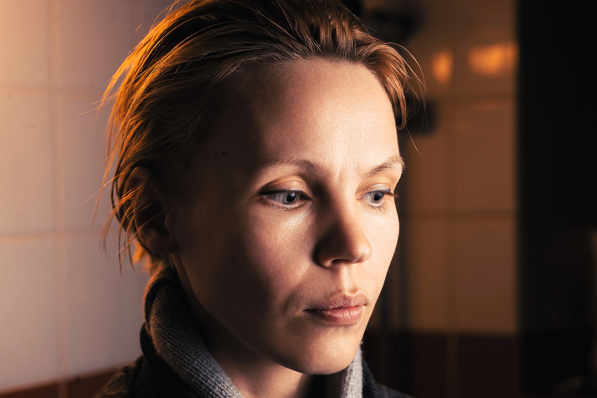 A person with short, light brown hair and light skin looks downward with a serious expression. Warm lighting illuminates their face, while a tiled wall is visible in the softly blurred background.