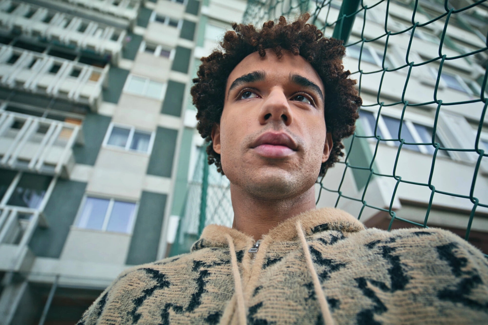 A young man with curly hair wearing a patterned hoodie stands in front of a tall apartment building, looking thoughtful. A green chain-link fence is visible beside him. The photo is taken from a low angle.