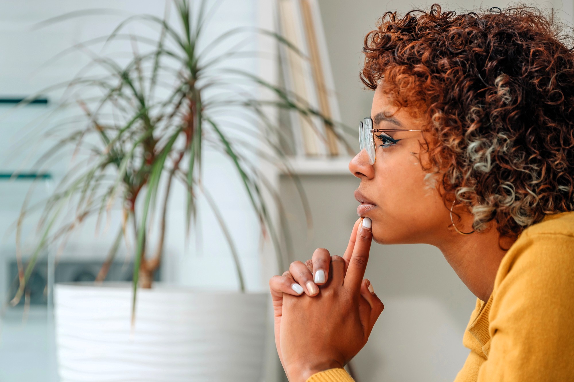 A person with curly hair and glasses sits in profile, resting their chin on their hand and appearing thoughtful. A large potted plant is in the background.