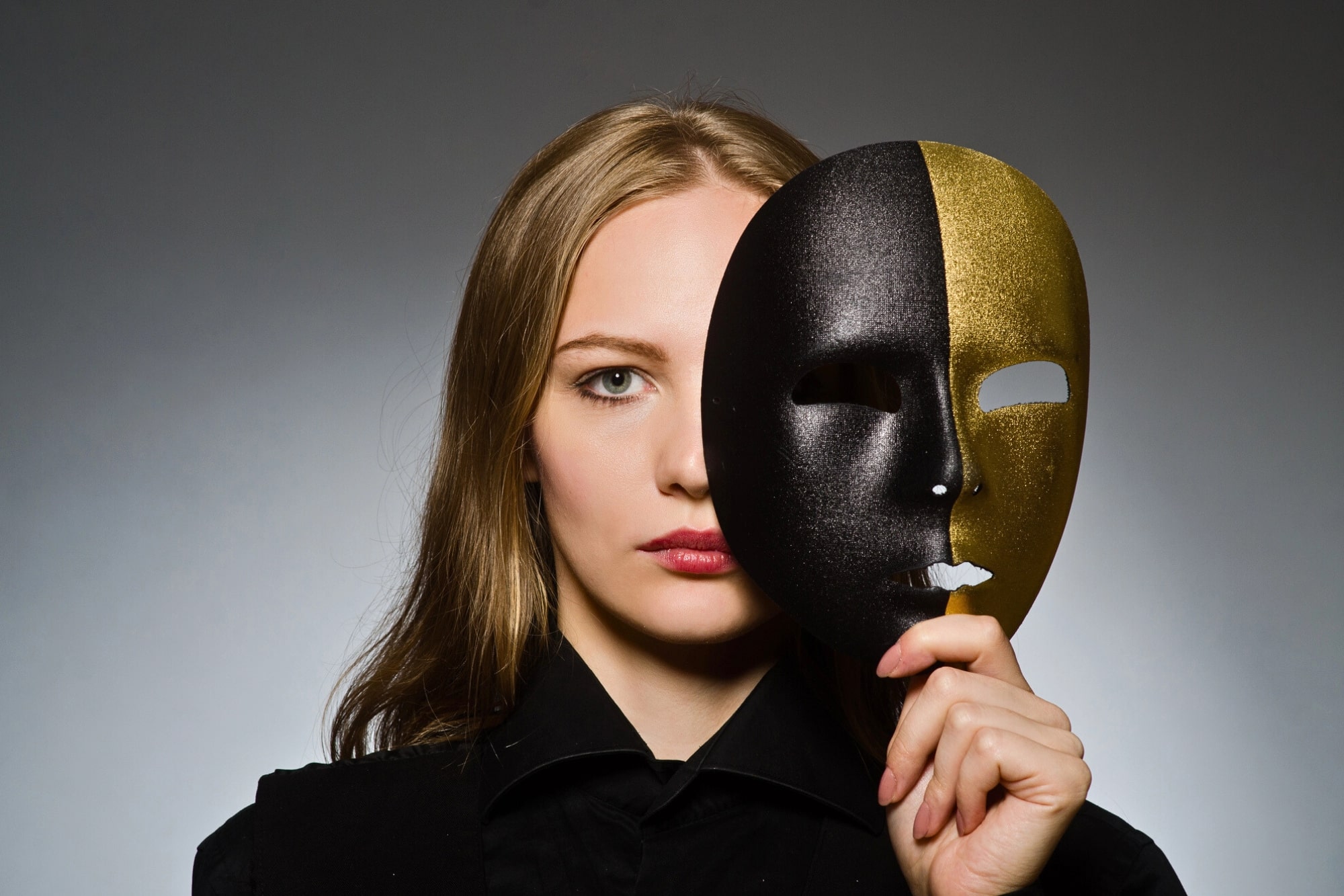 A woman with long blond hair holds a black and gold half-mask in front of her face, covering one eye, while the other half of her face is visible. She has a serious expression and stands against a gray background.