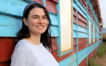 A woman with dark hair, wearing a light gray shirt and a headband, smiles while leaning against a weathered building painted in blue and red stripes. The background shows windows and a grassy area.