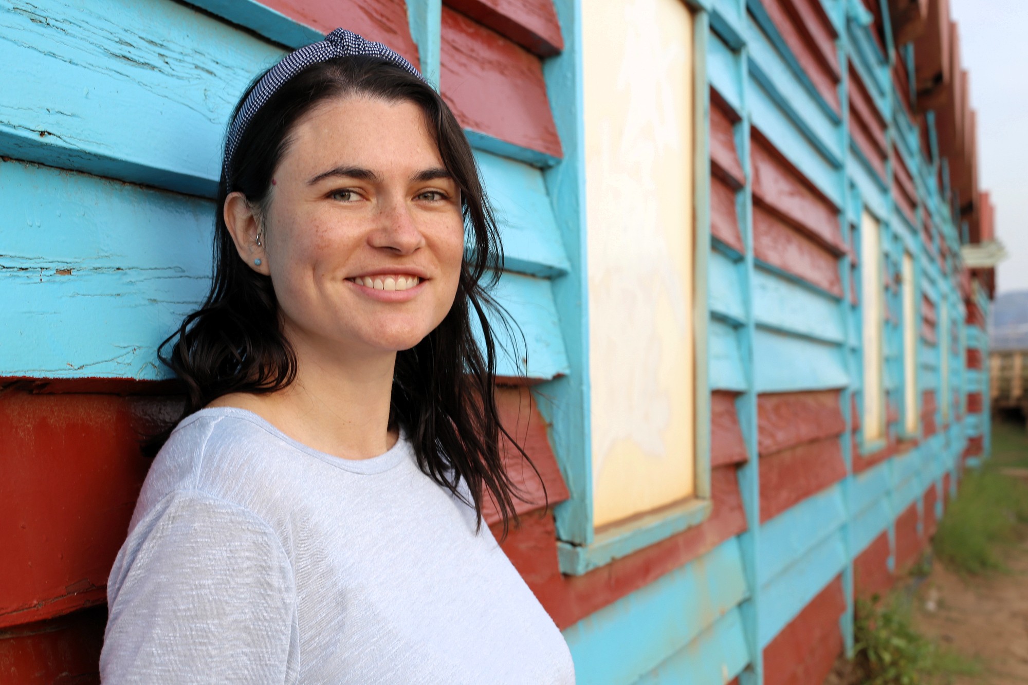 A woman with dark hair, wearing a light gray shirt and a headband, smiles while leaning against a weathered building painted in blue and red stripes. The background shows windows and a grassy area.