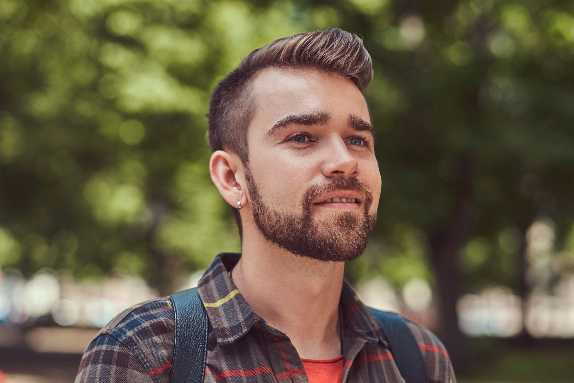 A young man with a beard and short hair, wearing a plaid shirt and backpack, stands outdoors with green trees blurred in the background. He is looking slightly upward and smiling softly.