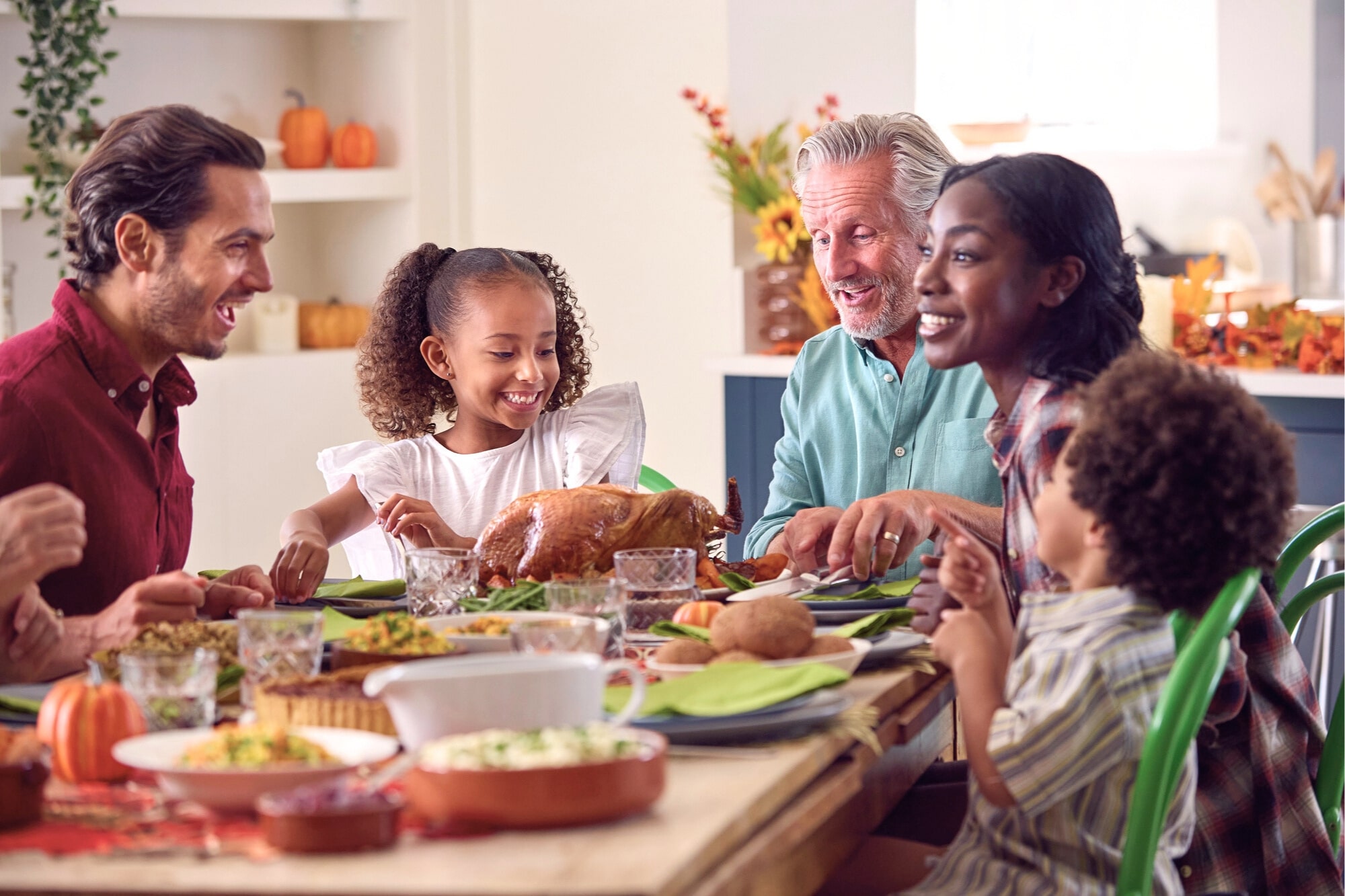 A diverse family of five joyfully shares a Thanksgiving meal at a decorated dining table, featuring a roast turkey, vegetables, and autumn-themed decor in a bright kitchen.