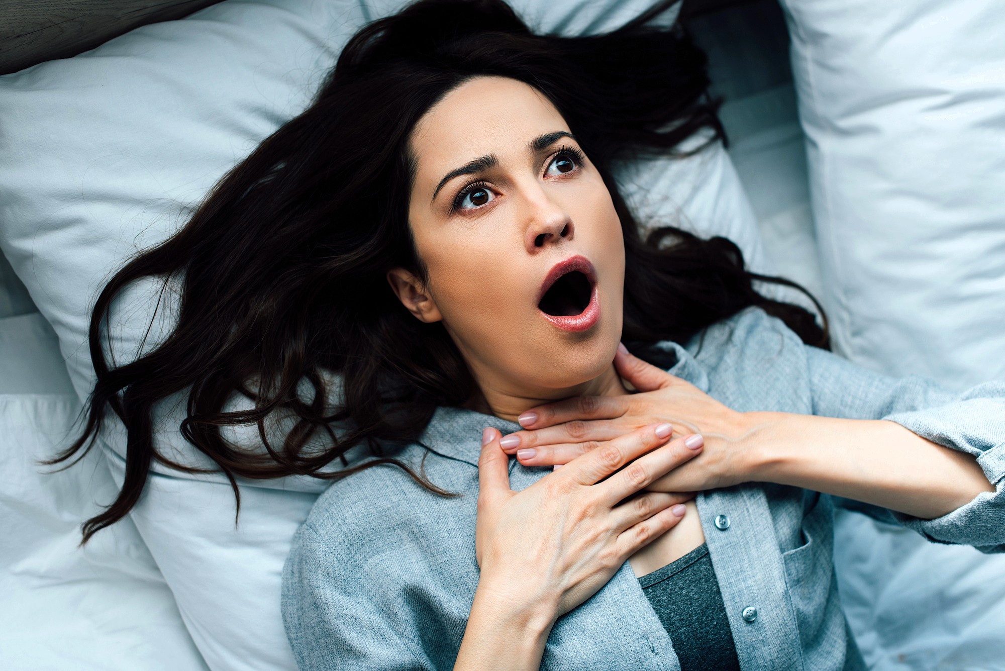 A woman lying on a bed looks alarmed, with wide eyes and mouth open, holding her hands to her throat as if struggling to breathe or feeling scared.