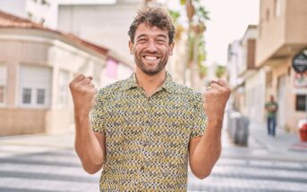 A young man with curly hair wearing a patterned shirt stands on a sunny city street, smiling broadly and raising both fists in a celebratory gesture.