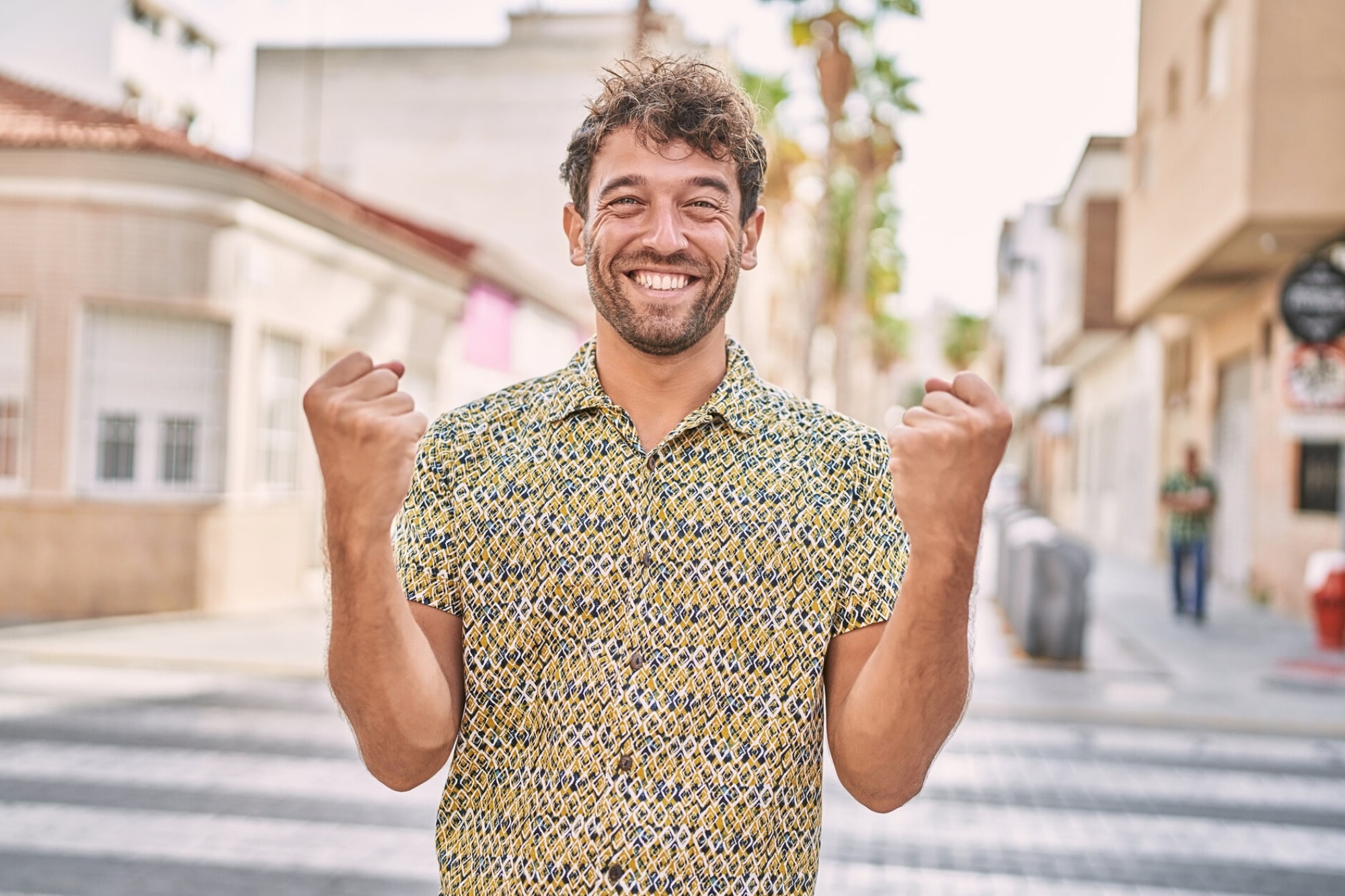 A young man with curly hair wearing a patterned shirt stands on a sunny city street, smiling broadly and raising both fists in a celebratory gesture.