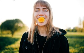 A young person with straight blonde hair stands outdoors, smiling with a yellow dandelion flower held in their mouth. They are wearing a black jacket, and greenery is visible in the blurred background.