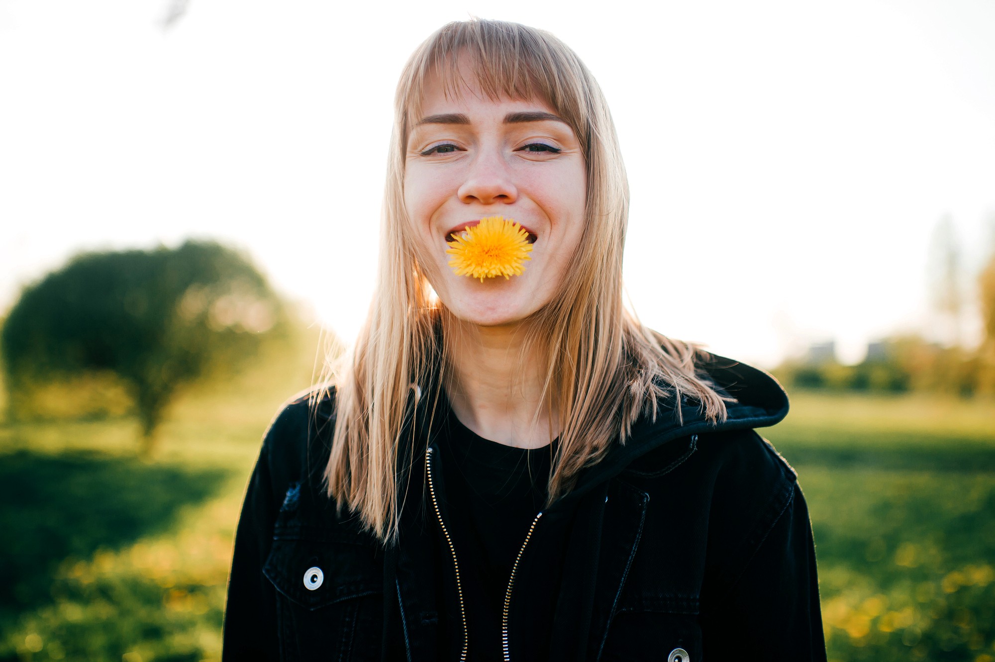 A young person with straight blonde hair stands outdoors, smiling with a yellow dandelion flower held in their mouth. They are wearing a black jacket, and greenery is visible in the blurred background.
