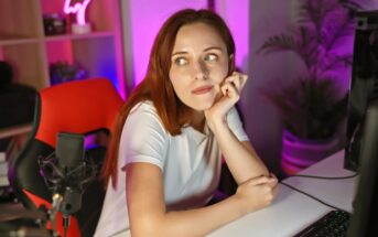 A young woman with long red hair, wearing a white shirt, sits at a desk with her chin resting on her hand, looking thoughtfully to the side in a colorful, softly lit room with a computer and microphone.