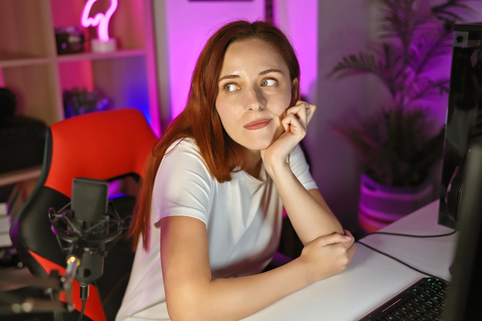 A young woman with long red hair, wearing a white shirt, sits at a desk with her chin resting on her hand, looking thoughtfully to the side in a colorful, softly lit room with a computer and microphone.