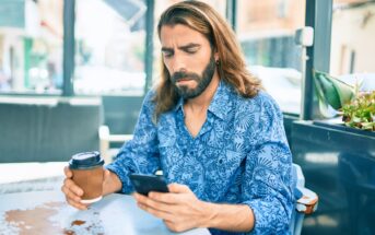 A man with long hair and a beard, wearing a blue patterned shirt, sits at a table in a cafe holding a coffee cup and looking at his smartphone with a serious expression.