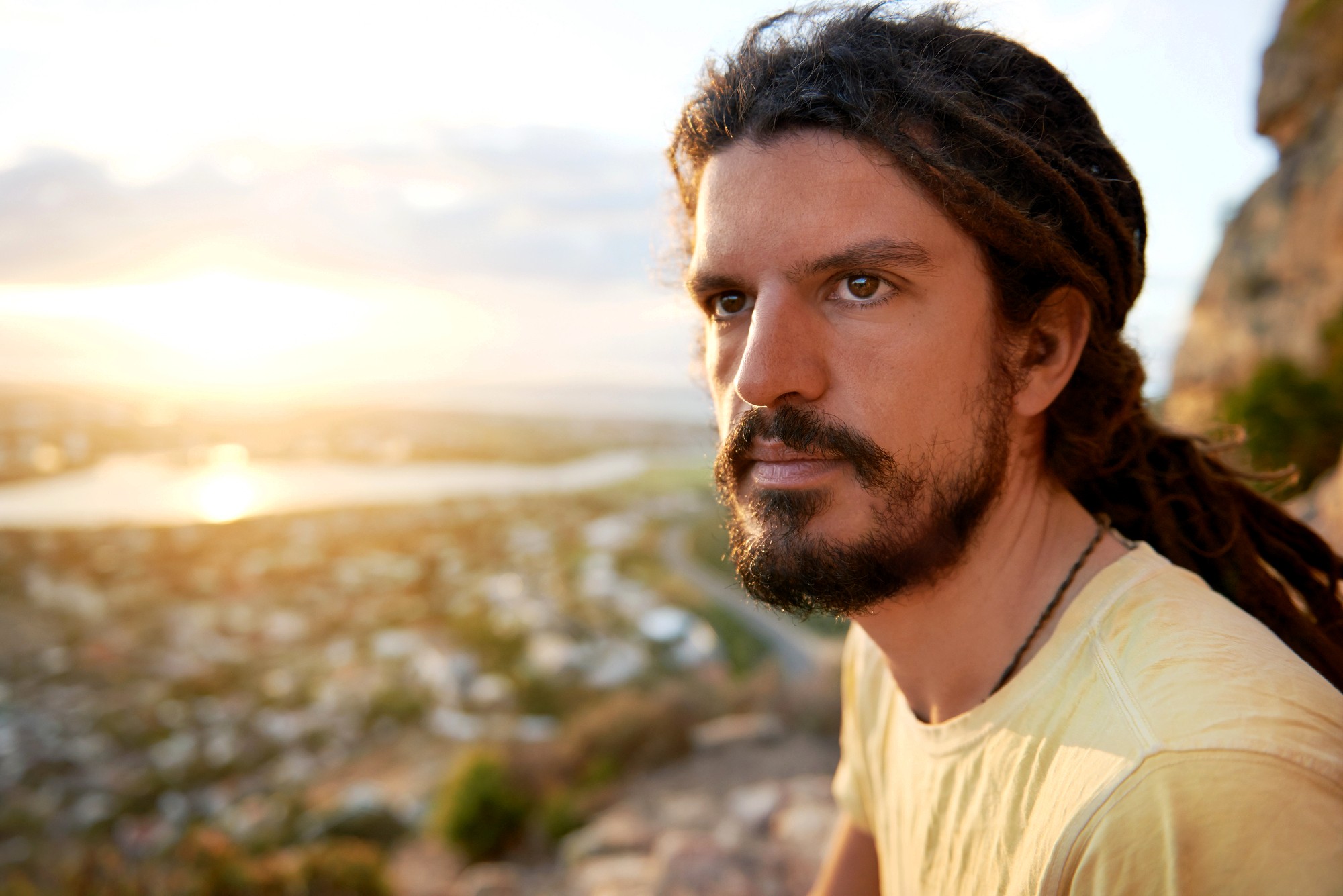 A man with long brown dreadlocks and a beard gazes thoughtfully into the distance, sitting outdoors with a blurred view of a sunlit landscape, including water and hills, in the background.