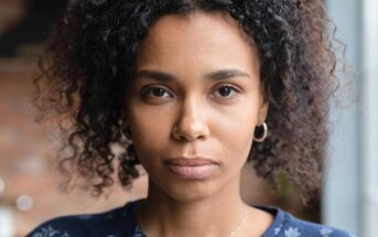 A woman with curly dark hair and hoop earrings looks directly at the camera with a neutral expression, wearing a blue top with a subtle floral pattern. The background is softly blurred.