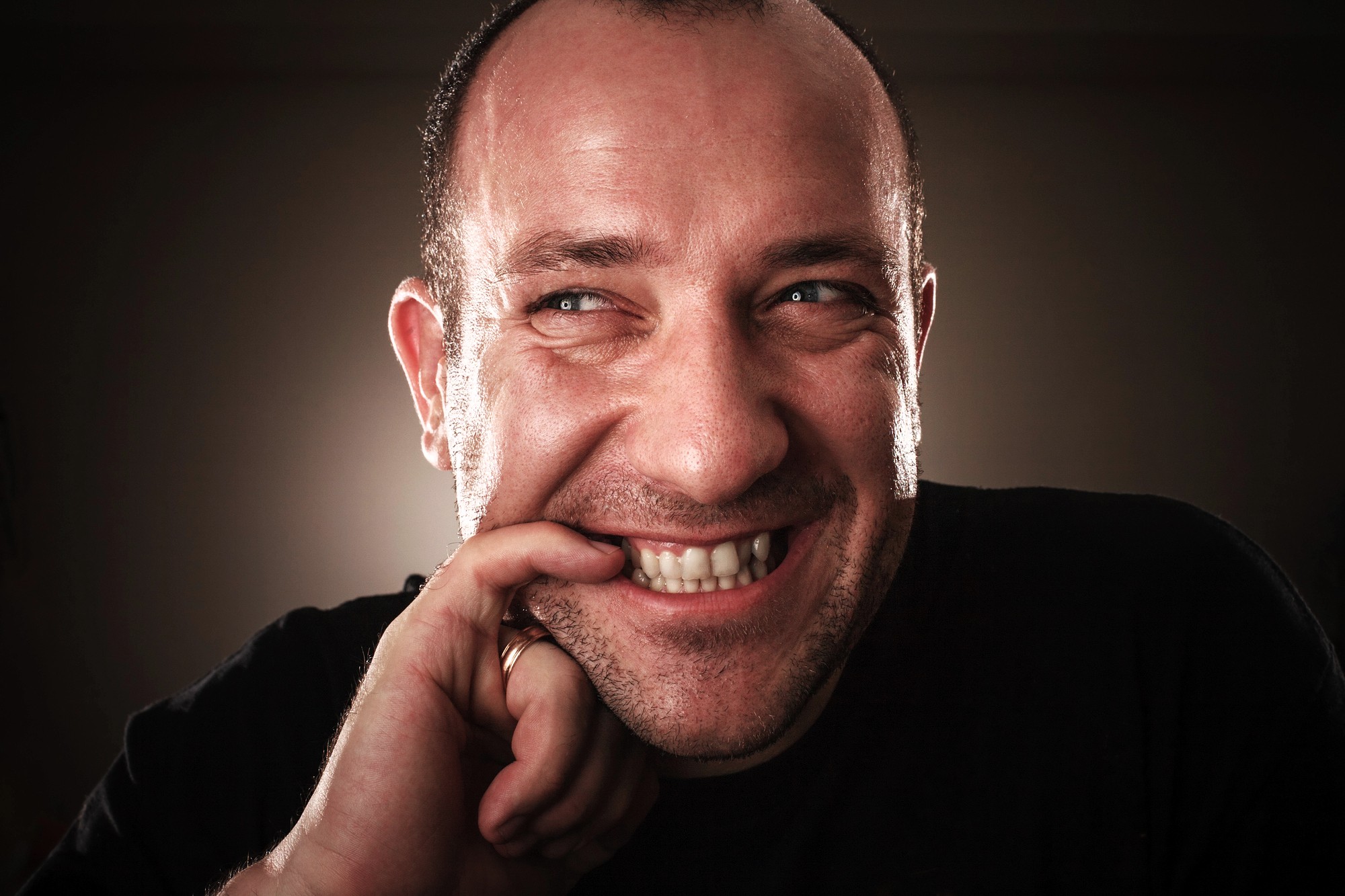 A man with light skin smiles playfully, biting his index finger. He is looking up and to the side, with a mischievous expression. The background is dark, and he is wearing a black shirt.