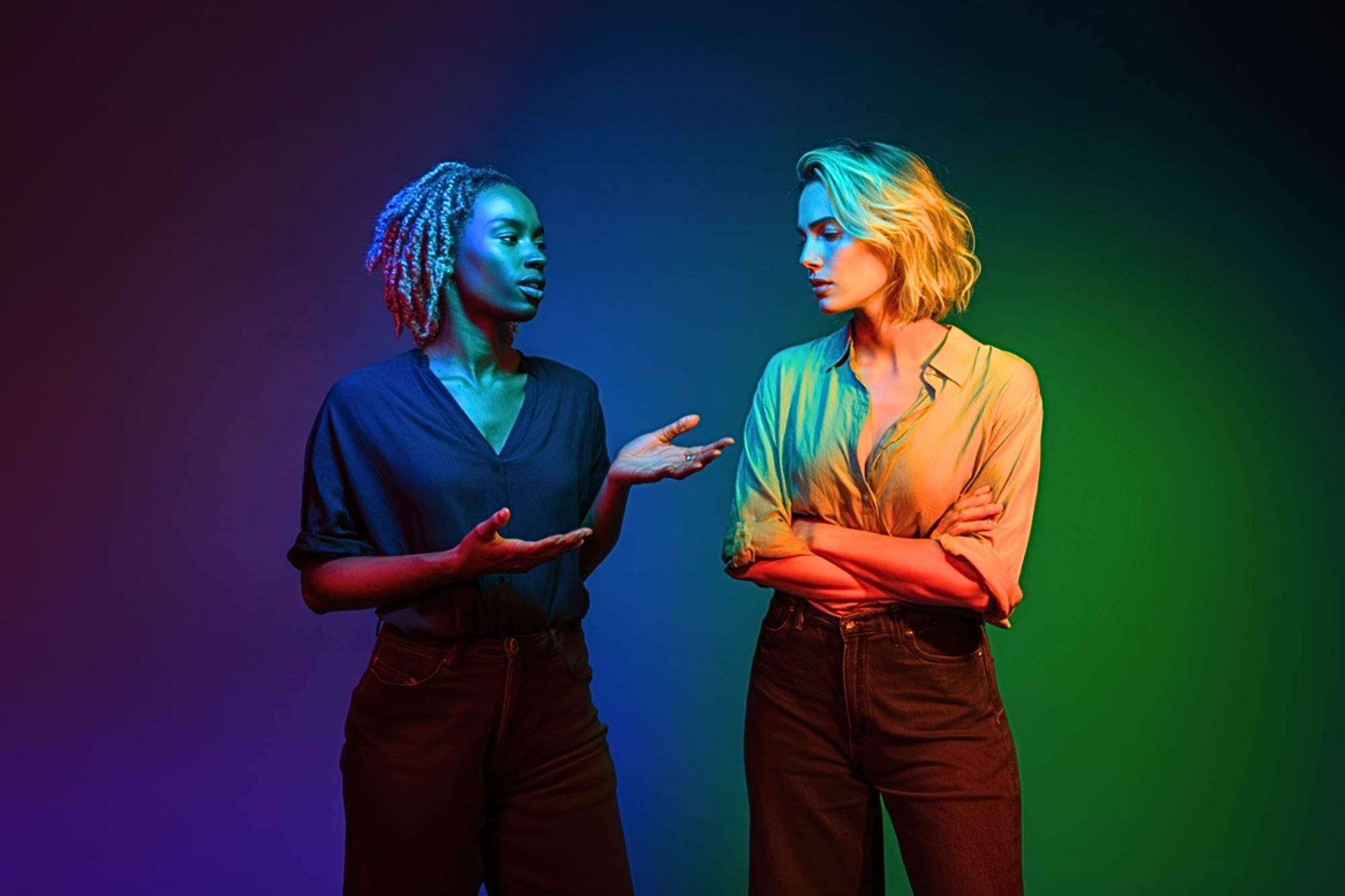 Two women stand facing each other under dramatic, colorful lighting. One gestures with her hands while speaking; the other stands with arms crossed, listening. The background blends blue, red, and green hues.