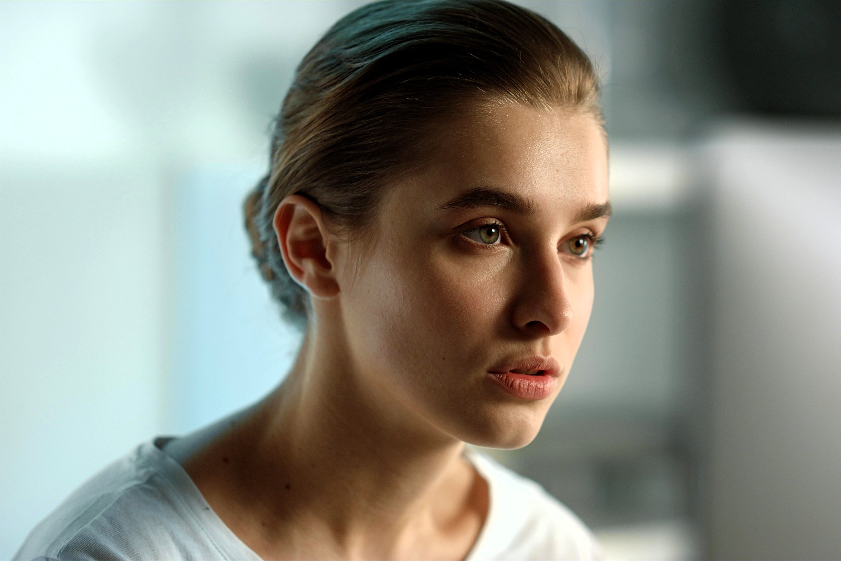 A young woman with light skin and brown hair pulled back looks thoughtfully ahead. She wears a white shirt and is in a softly lit, blurred indoor setting.
