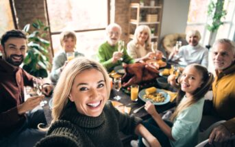 A group of people of various ages sit around a dinner table with a roast turkey, smiling and raising glasses, while a woman in front takes a selfie. The scene appears cheerful and festive.