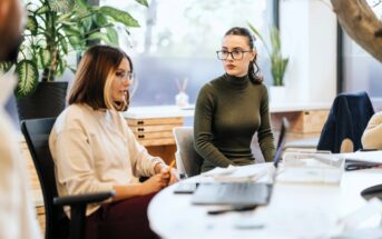 Two women sitting at a desk in a modern office, having a discussion. One is wearing a white shirt, the other a green turtleneck. Papers, a laptop, and office supplies are on the table, with plants and large windows in the background.