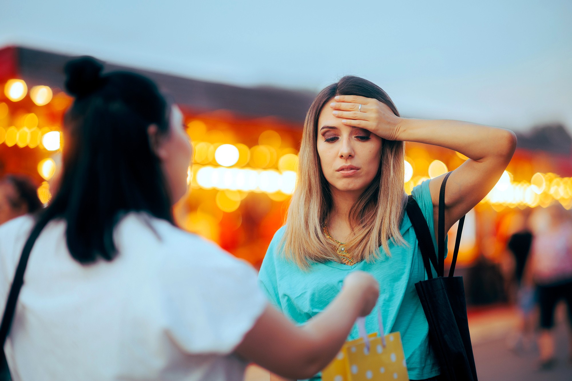 A woman touches her forehead and looks unwell while another person hands her a shopping bag at an outdoor market with blurred lights in the background.