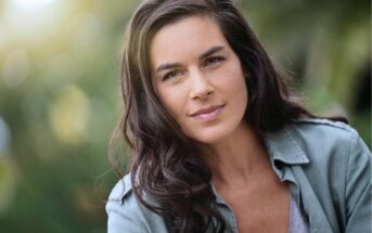 A woman with long, dark hair wearing a light green button-up shirt looks confidently at the camera. The background is blurred greenery, giving the image a natural, outdoor feel.