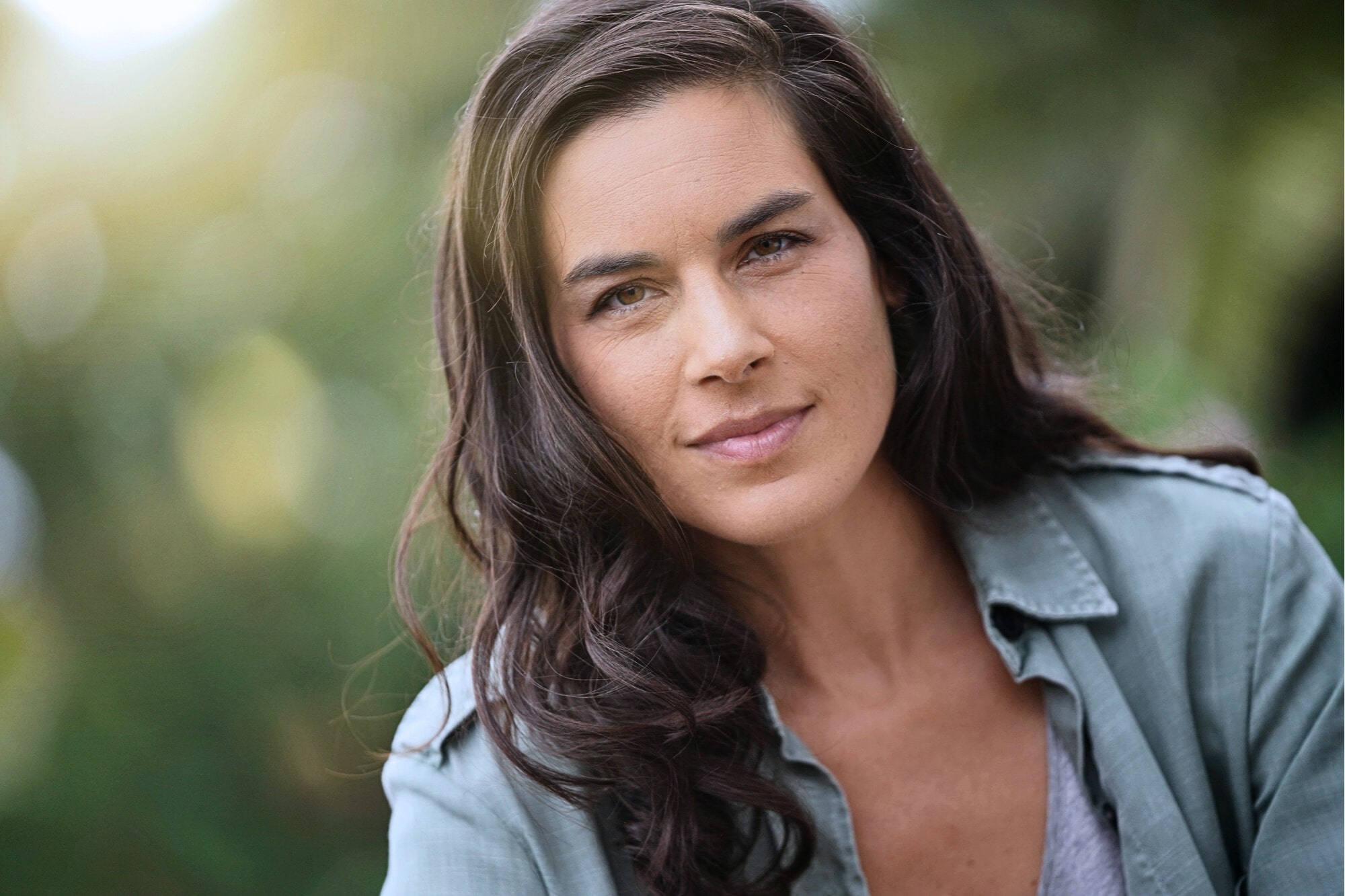 A woman with long, dark hair wearing a light green button-up shirt looks confidently at the camera. The background is blurred greenery, giving the image a natural, outdoor feel.