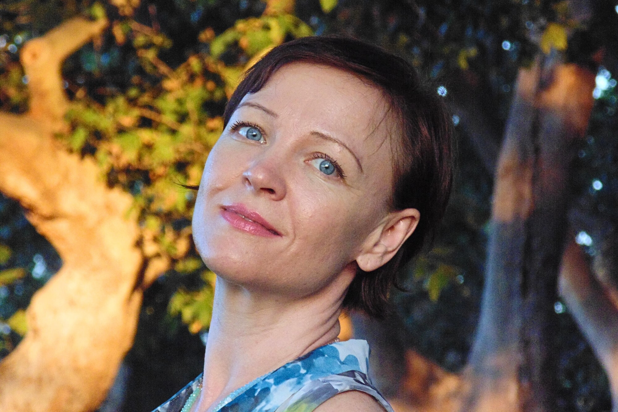 A woman with short brown hair and blue eyes smiles slightly, standing outdoors with sunlight and trees in the background. She is wearing a blue and white patterned sleeveless top.