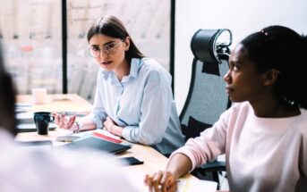Two women sit at a conference table in a modern office, engaged in discussion. One woman with glasses speaks, while the other listens attentively. Notebooks, documents, and a coffee mug are on the table.