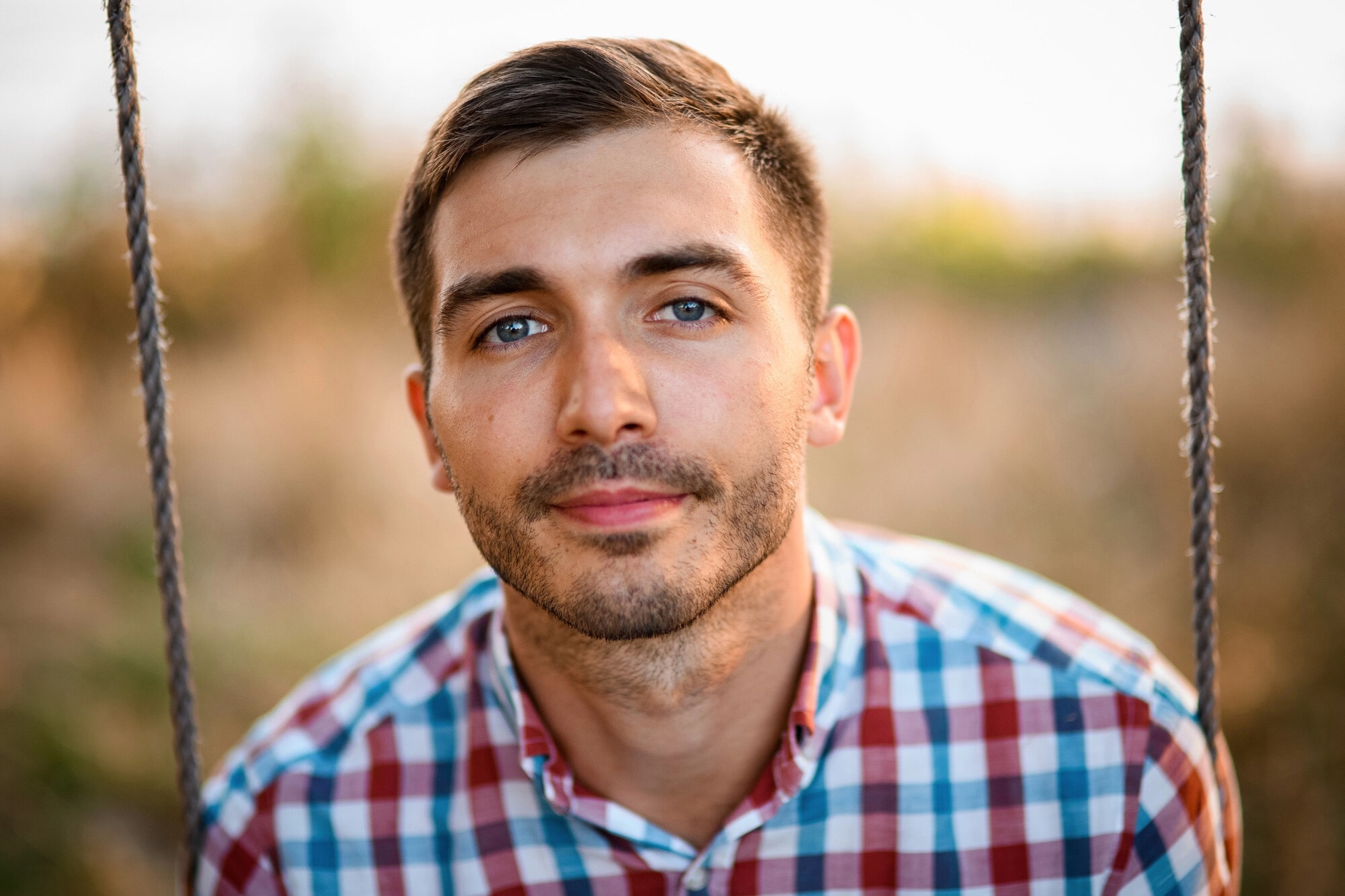 A young man with short brown hair and blue eyes, wearing a red, blue, and white plaid shirt, sits on a swing outdoors, looking directly at the camera, with a blurred natural background.