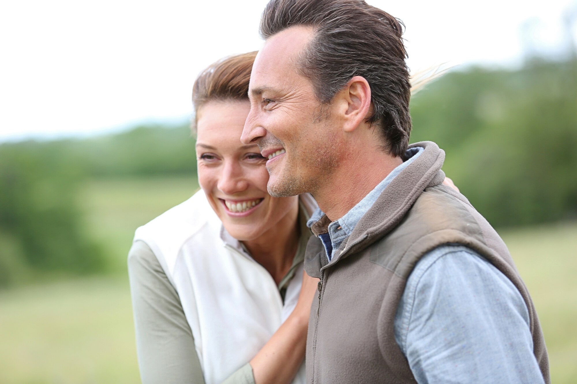 A smiling couple stands close together outdoors, with the woman resting her hand on the man's shoulder. They both appear happy and are dressed in casual outdoor clothing, with a blurred green landscape in the background.