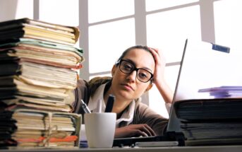 A woman wearing glasses sits at a desk with a large stack of folders and papers, looking tired. A coffee mug, pens, and a laptop are also on the desk, and sunlight streams in through the window behind her.