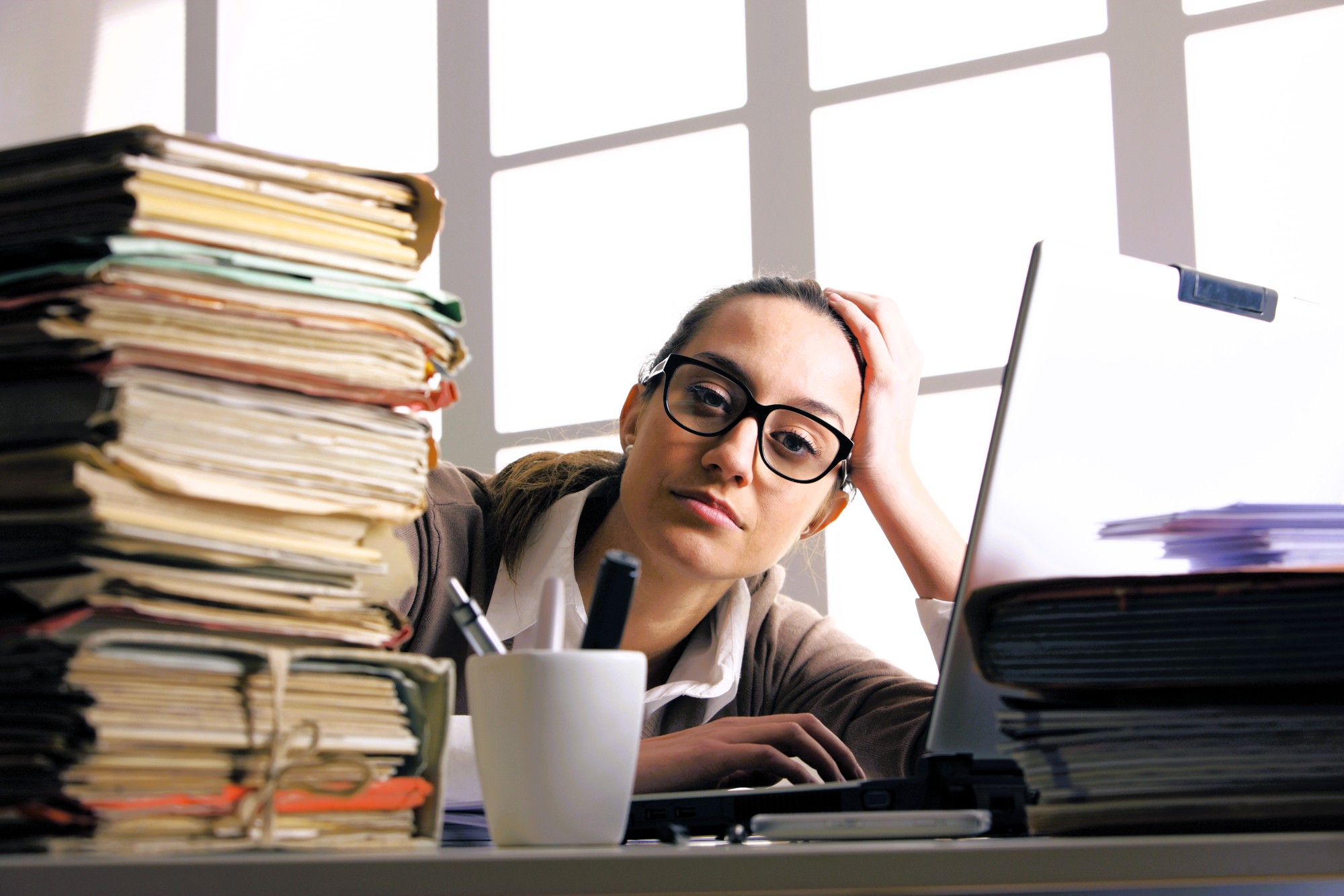 A woman wearing glasses sits at a desk with a large stack of folders and papers, looking tired. A coffee mug, pens, and a laptop are also on the desk, and sunlight streams in through the window behind her.