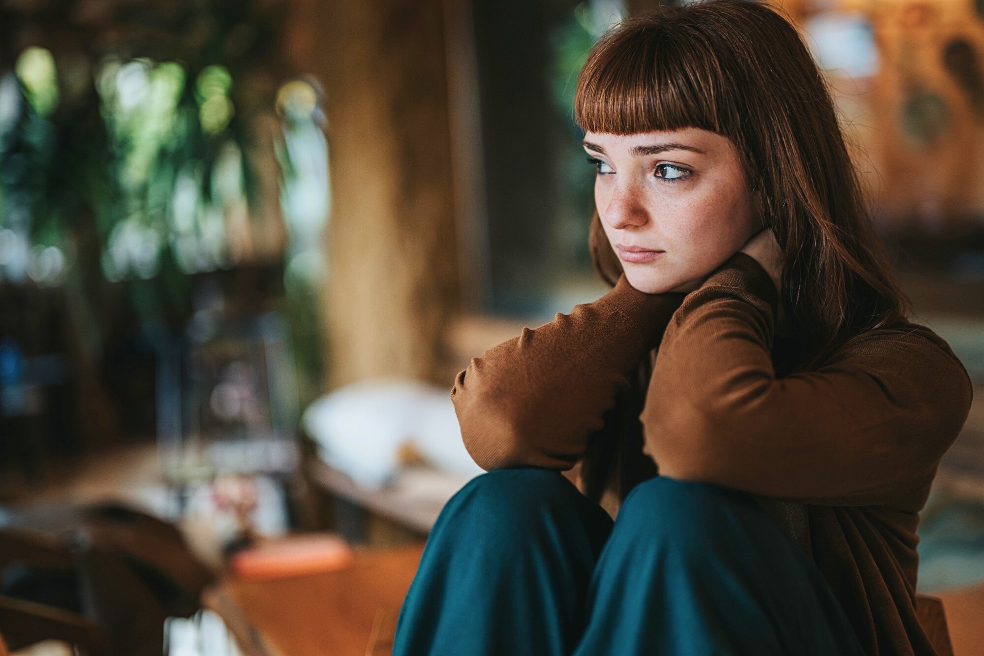A young woman with straight brown hair and bangs sits indoors, hugging her knees and looking pensively to the side. She wears a brown top and green pants. The background is softly blurred with warm light and plants.