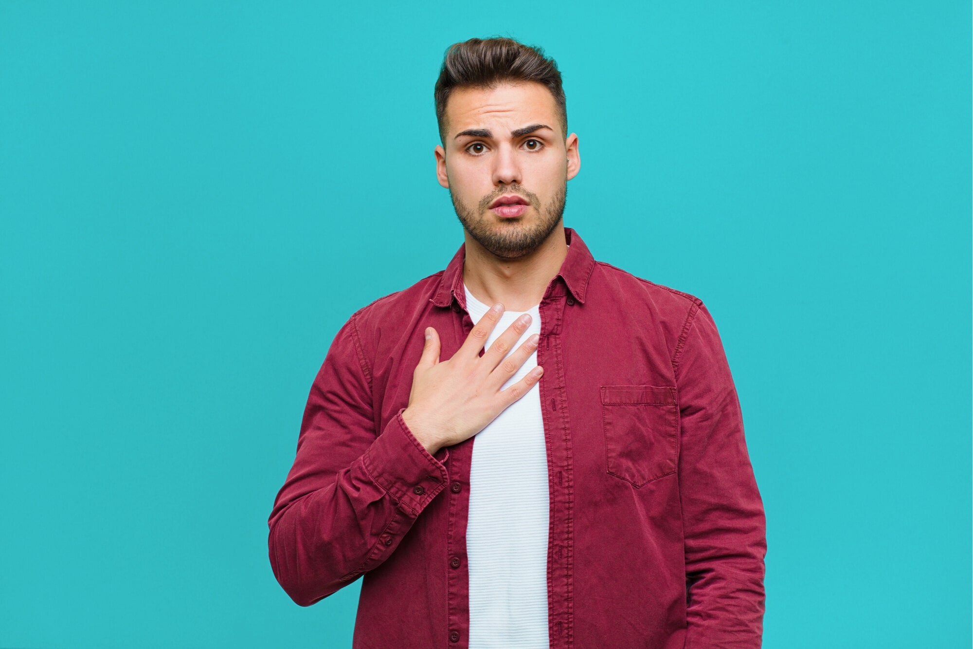 A young man with short dark hair, wearing a maroon button-up shirt over a white t-shirt, stands against a teal background, looking surprised and touching his chest with one hand.