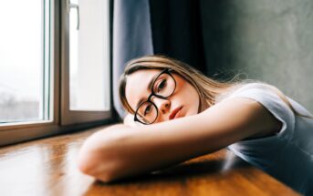 A young woman wearing glasses rests her head and arm on a wooden table next to a window, looking thoughtfully outside. Soft natural light illuminates her face and the room appears calm and quiet.