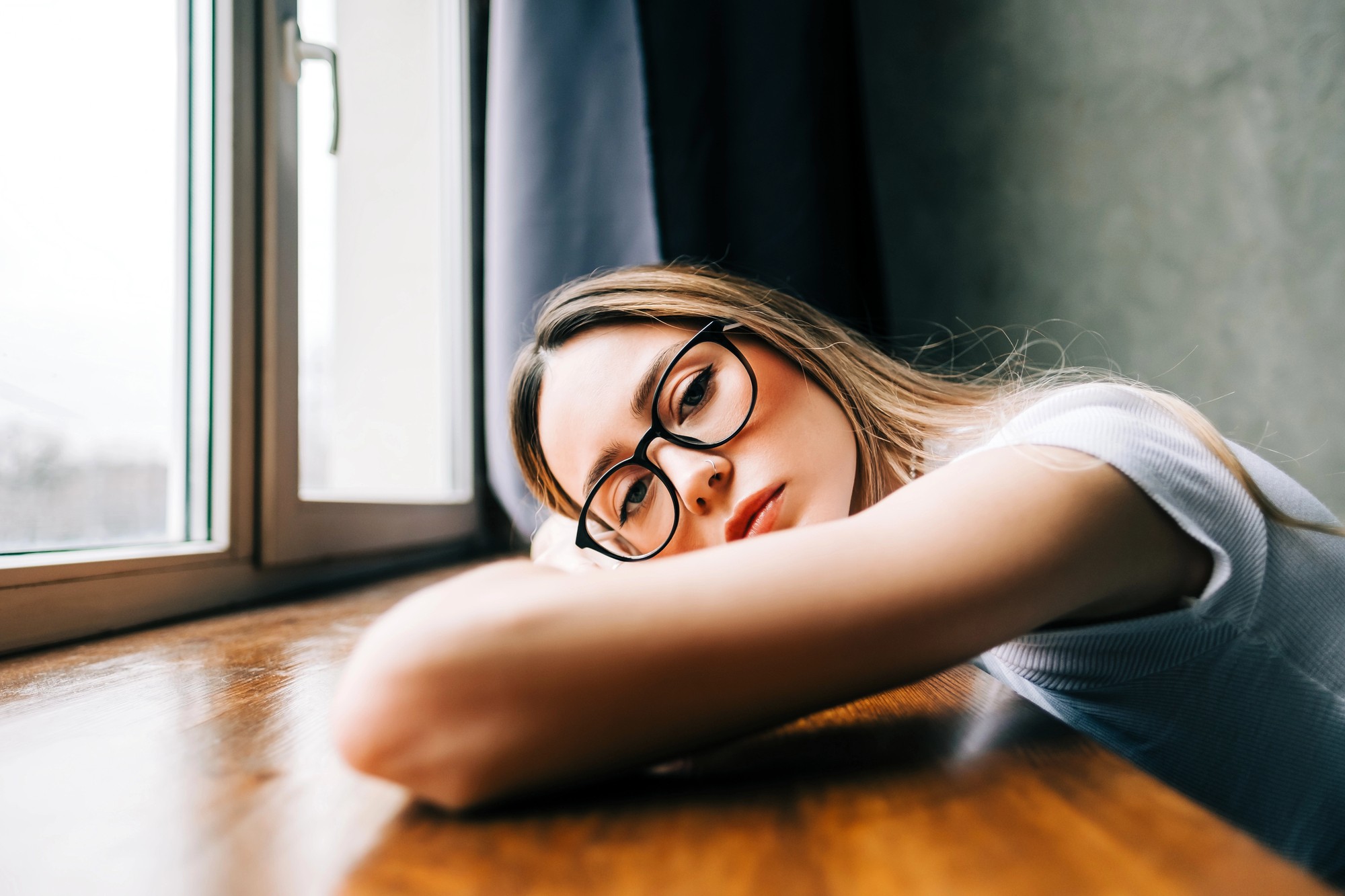 A young woman wearing glasses rests her head and arm on a wooden table next to a window, looking thoughtfully outside. Soft natural light illuminates her face and the room appears calm and quiet.