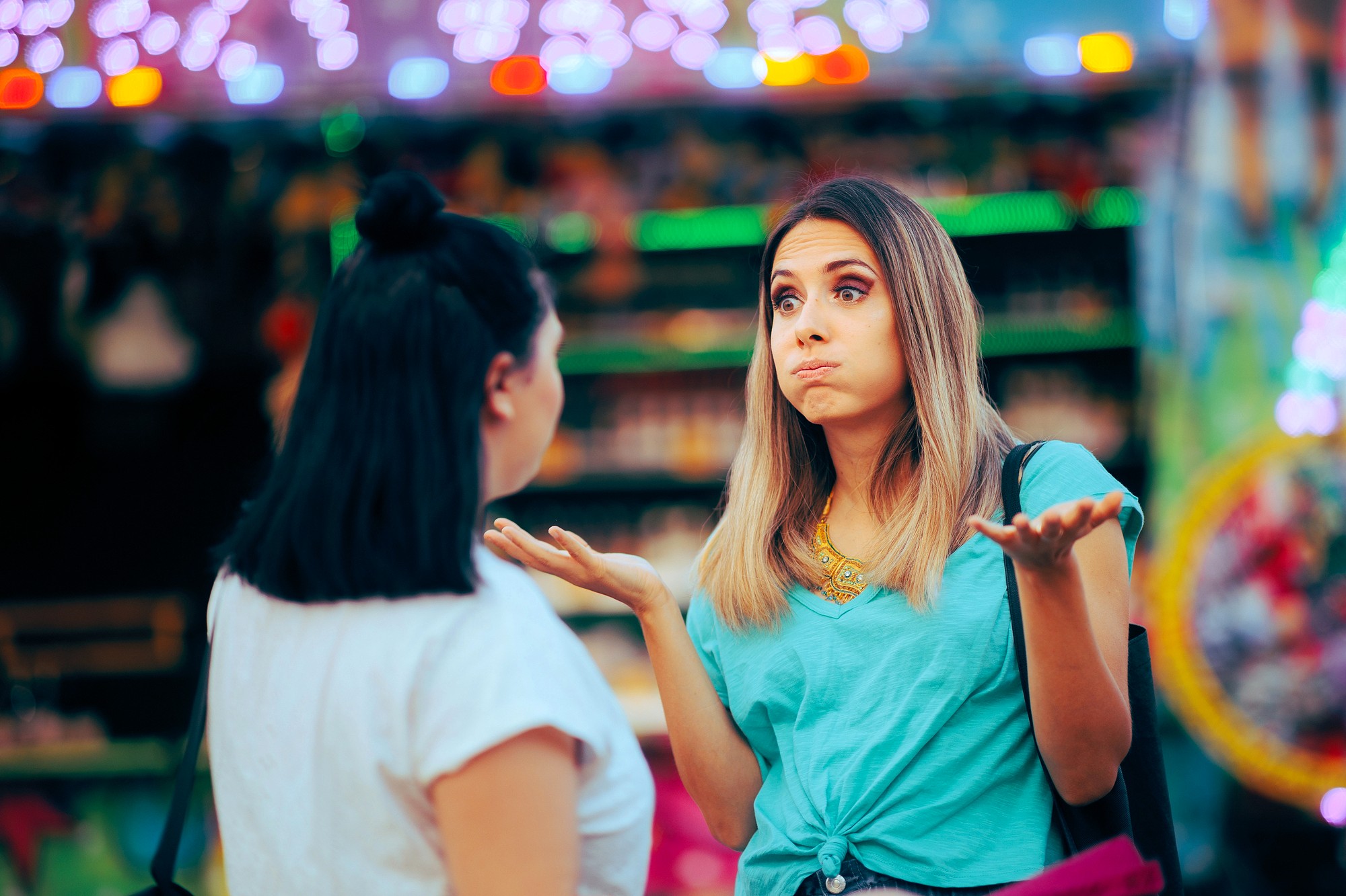 Two women are talking outdoors; one woman with long hair shrugs with her hands up and cheeks puffed out, appearing confused or unsure, while the other woman with dark hair listens. Colorful lights are blurred in the background.
