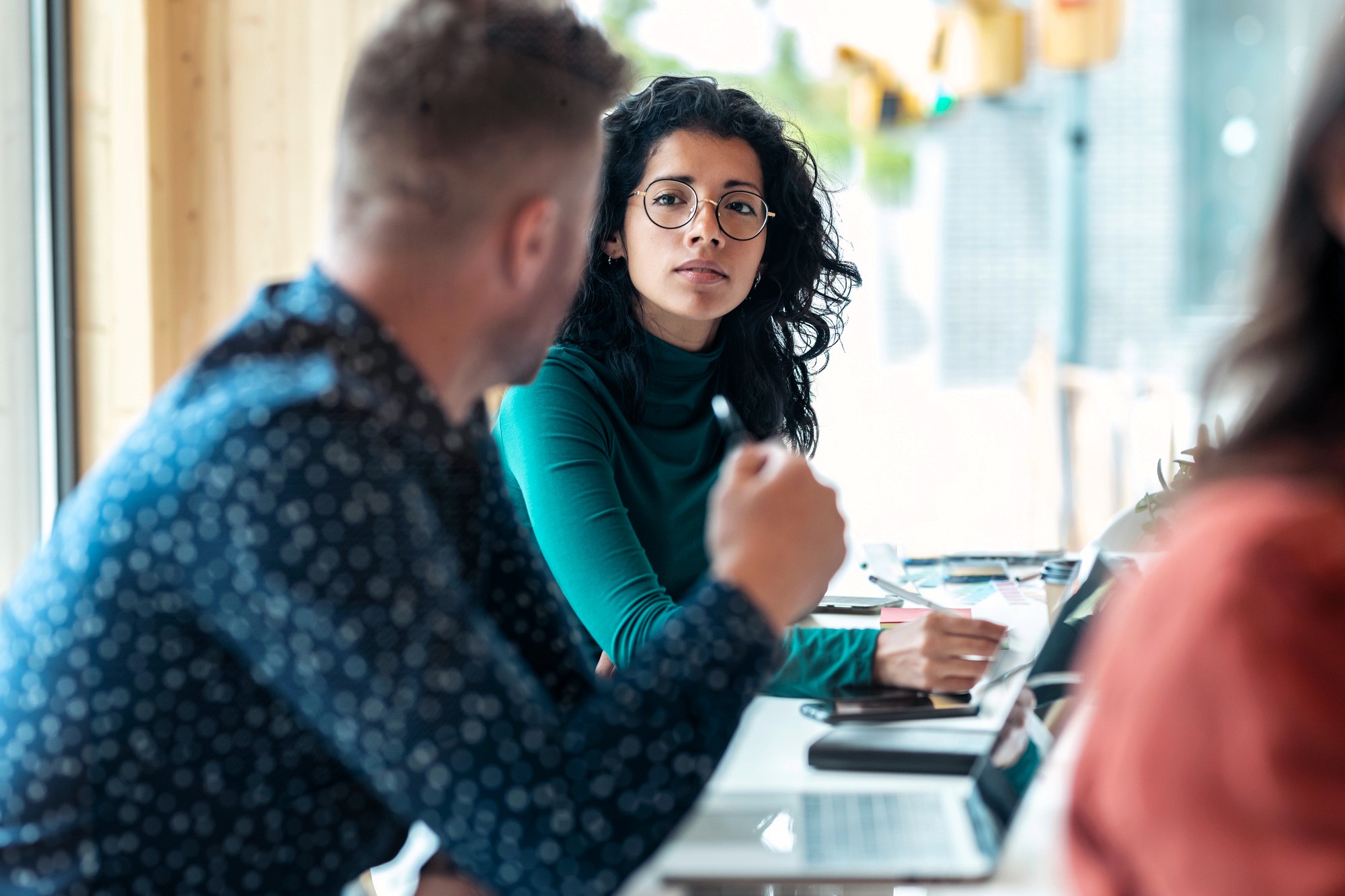 A woman with curly hair and glasses sits at a table, looking attentively at a man who is speaking. They appear to be in a discussion, possibly in a casual office or café setting, with notebooks and laptops on the table.