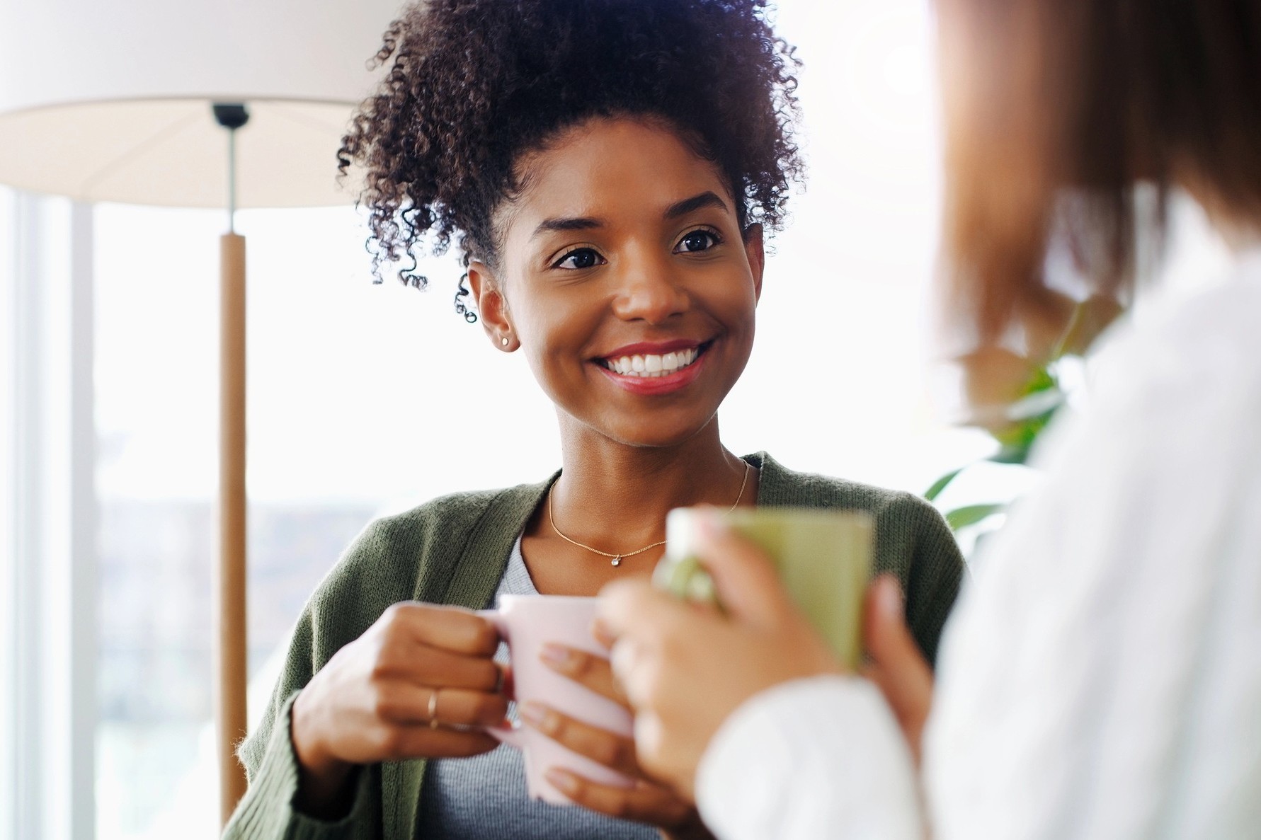 Two women sit together indoors, holding mugs and smiling warmly at each other, suggesting a friendly conversation in a cozy, well-lit room with a lamp and plants in the background.