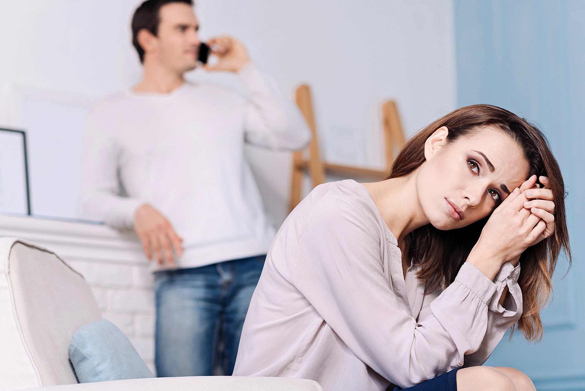A woman sits on a couch looking upset, resting her head on her hands. In the background, a man stands and talks on a phone, appearing indifferent or distracted. The scene suggests tension or emotional distance.