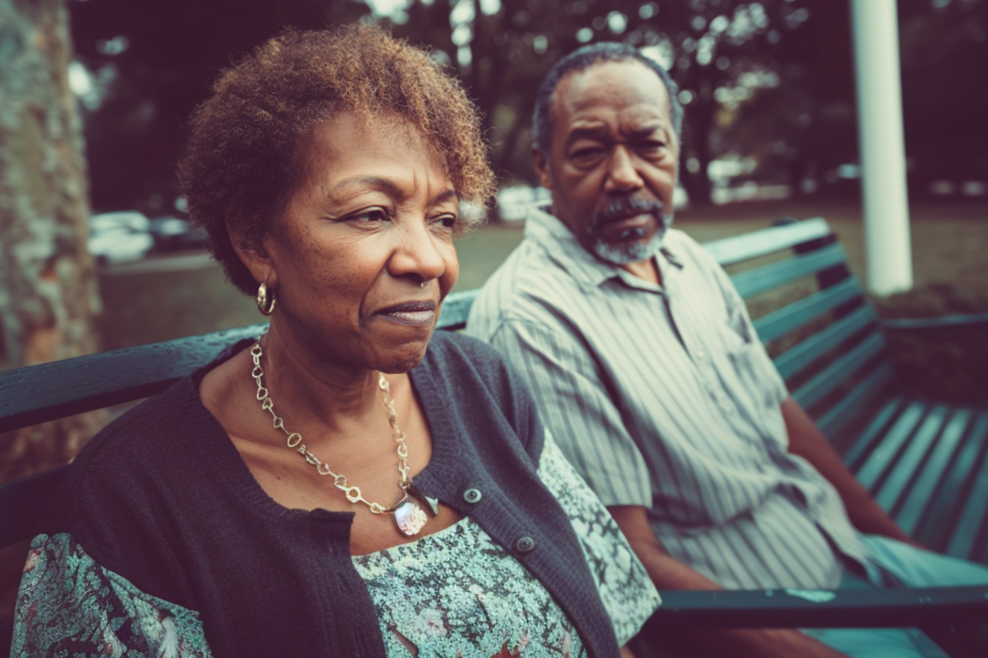 An older woman and man sit on a green park bench. The woman looks thoughtful or concerned, while the man gazes toward her. Trees and a grassy area are visible in the background.