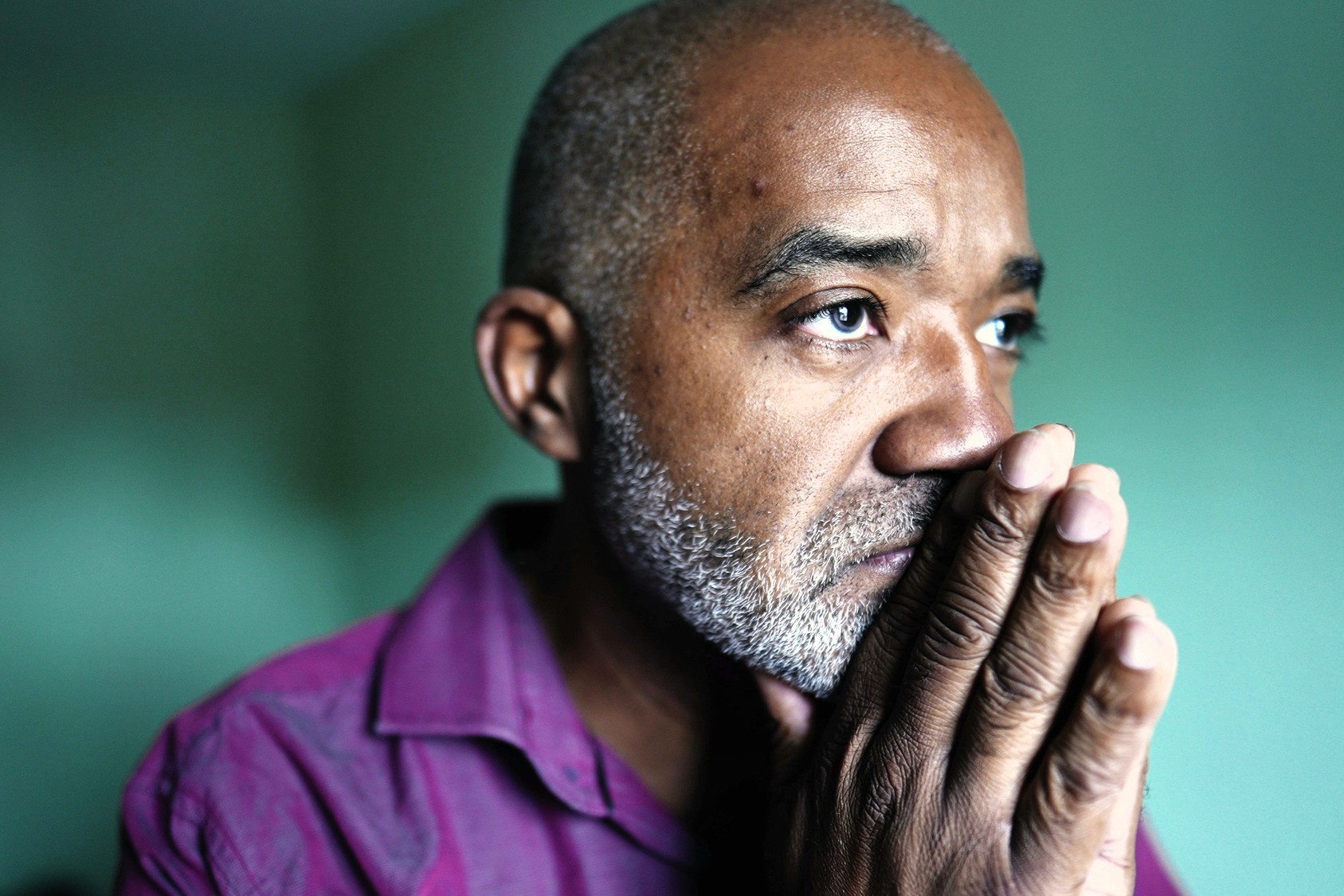 A middle-aged man with a short beard and mustache looks thoughtful and concerned, resting his hands against his face. He is wearing a purple shirt and gazing into the distance in a softly lit room.