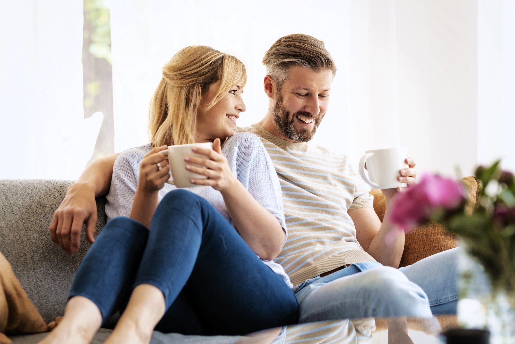 A smiling couple sits close together on a couch, each holding a mug. They appear relaxed and happy, looking at each other, with natural light coming from a window behind them and flowers blurred in the foreground.