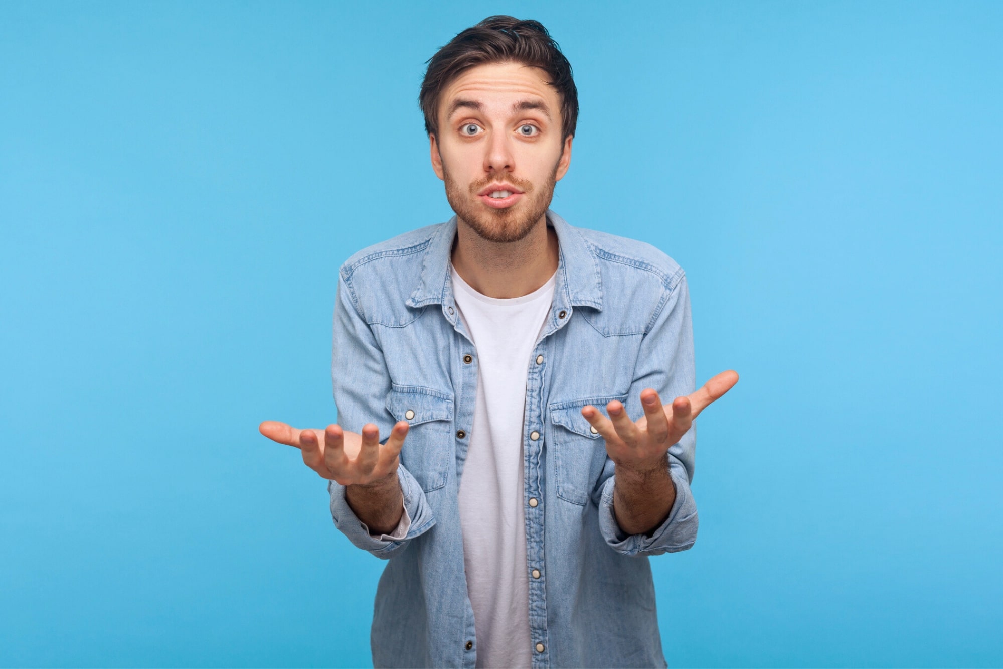 A young man in a light denim shirt and white t-shirt stands against a blue background, looking confused or questioning, with both hands raised and palms facing up.