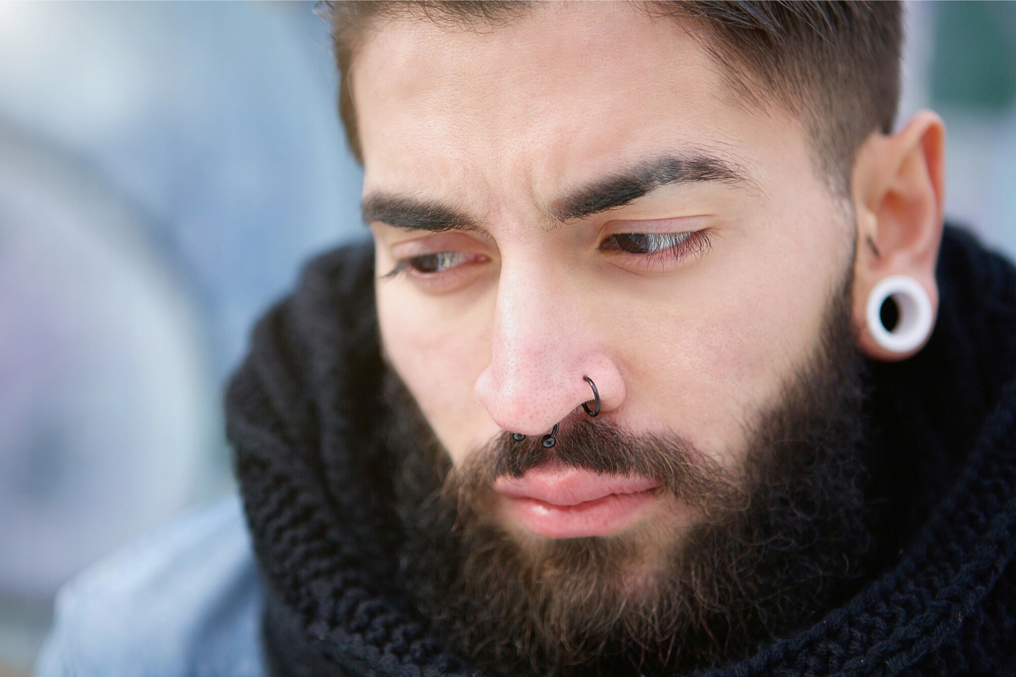 A close-up of a young man with a beard, dark hair, stretched earlobe piercings, and a nose ring, looking down with a serious or contemplative expression. He is wearing a black scarf and a blue jacket.