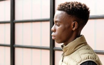 A young person with short, curly hair is shown in profile, wearing a light tan vest over a dark shirt, standing in front of a window with frosted glass panels.