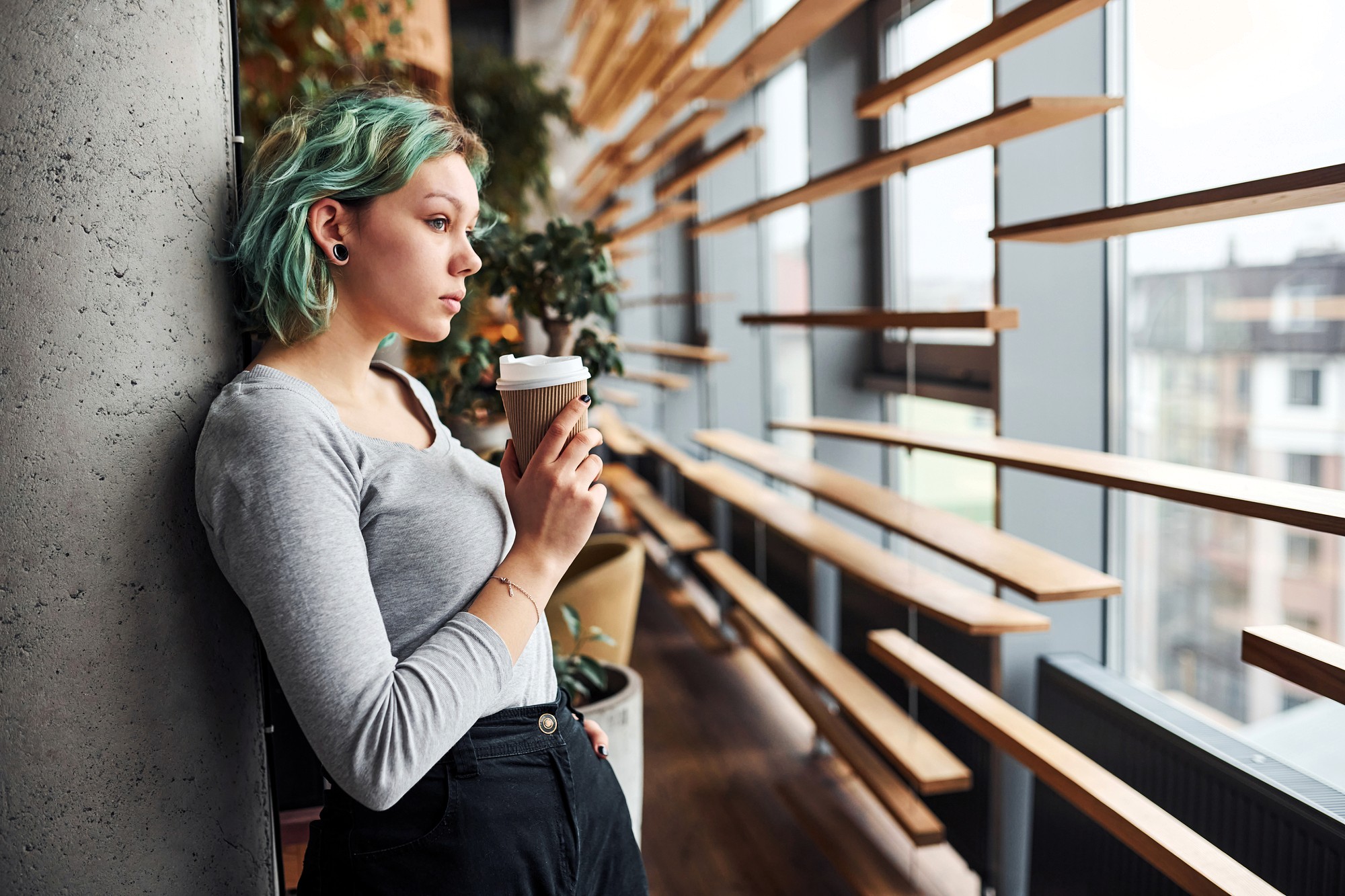 Young woman with short turquoise hair leans against a concrete wall, holding a takeaway coffee cup. She looks thoughtful, gazing out large windows in a modern, stylish space with wooden slats and indoor plants.