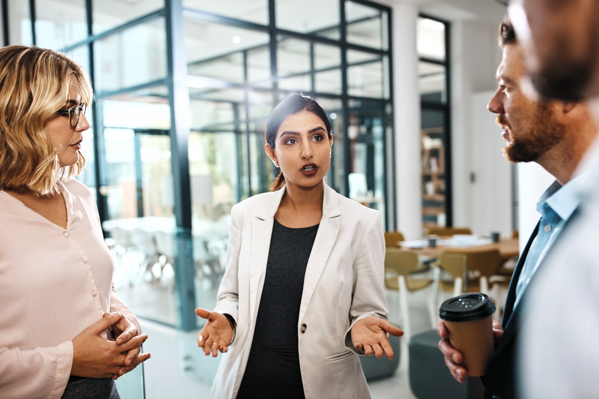 Four people stand in an office, engaged in conversation. One woman in a white blazer speaks while gesturing with her hands. Two others listen intently, and one person holds a coffee cup. Glass walls and tables are visible in the background.