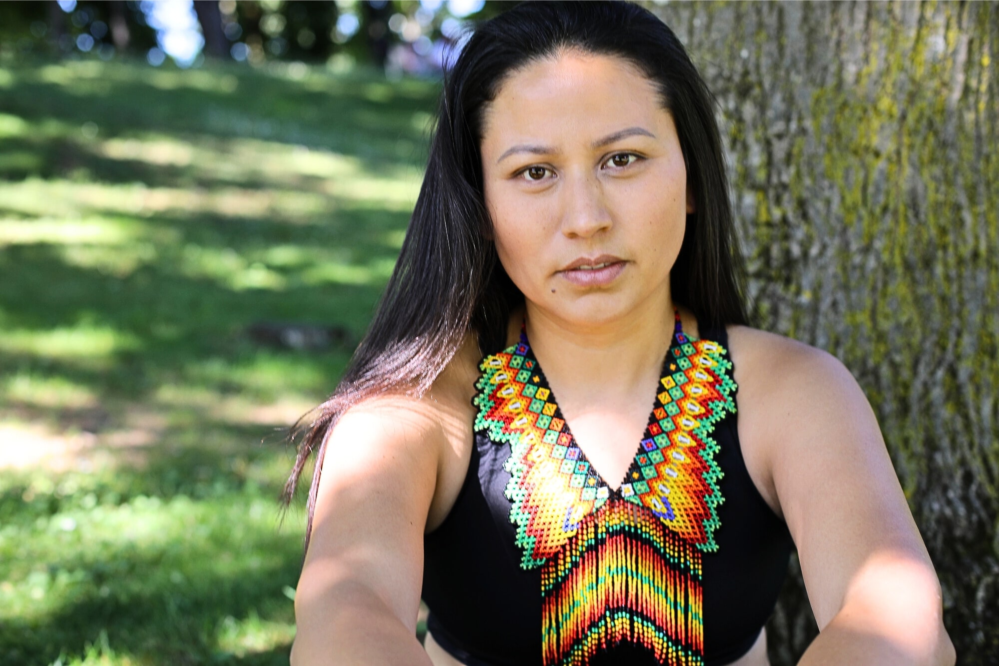A woman with long dark hair sits outdoors by a tree, wearing a black top and a colorful beaded necklace with geometric patterns. Sunlight filters through the trees onto the grass in the background.