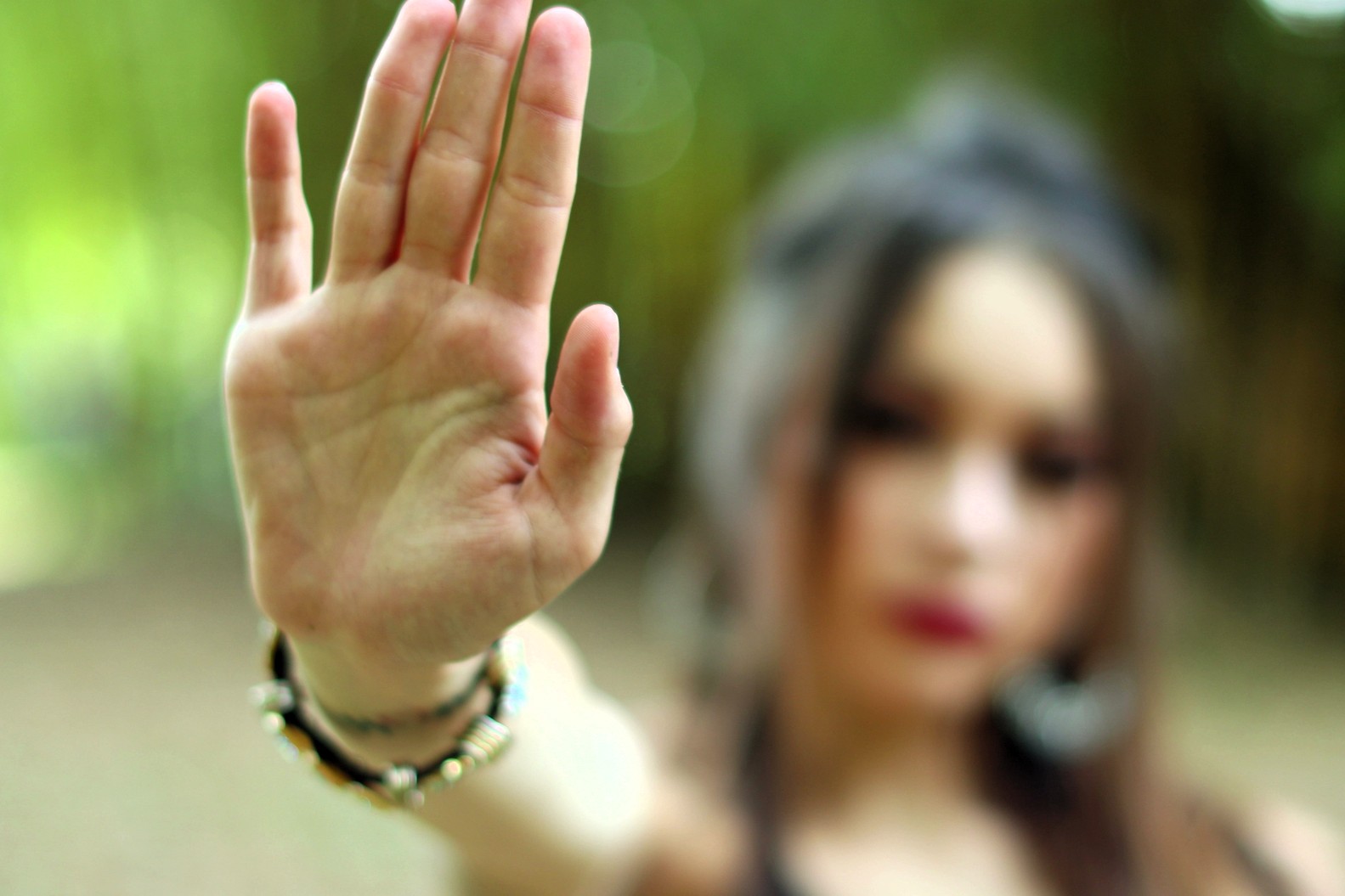 A person with long hair holds their hand up toward the camera, palm facing forward, signaling “stop.” The background is blurred and green, and the person’s face is out of focus.